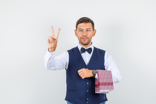 Serveur en chemise, gilet, noeud papillon montrant le signe de la victoire et à la vue de face, heureux.