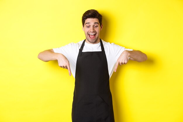 Serveur excité en tablier noir pointant les doigts vers le bas, vérifiant l'offre promotionnelle, debout sur fond jaune.