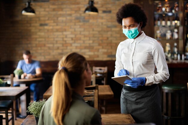 Serveuse afro-américaine avec masque facial et gants à l'aide d'une tablette numérique tout en prenant la commande d'un gues