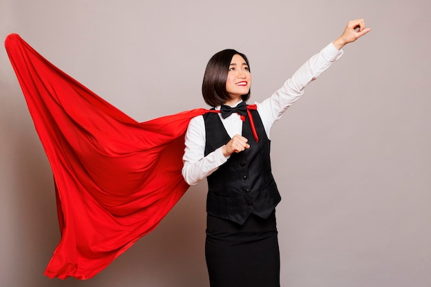 Photo gratuite une serveuse asiatique souriante, une super-femme en cape flottante, montrant sa force et sa puissance, une réceptionniste confiante et joyeuse portant la cape rouge de superman, levant le poing serré et levant les yeux.