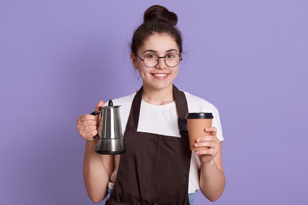 Serveuse souriante en t-shirt blanc et tablier marron tenant le pot et emporter la tasse dans les mains