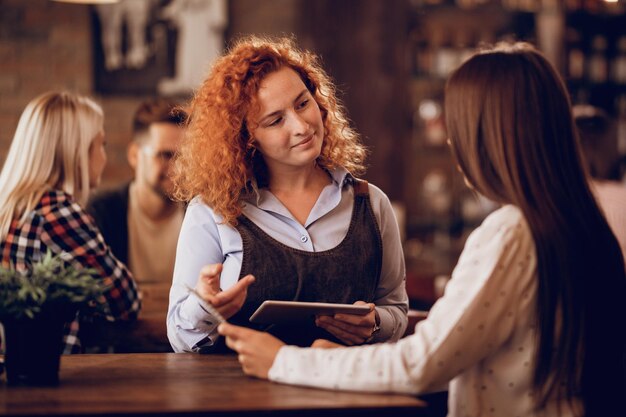 Serveuse souriante utilisant une tablette numérique tout en parlant à un invité et en prenant sa commande dans un pub