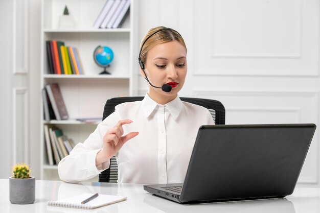 Service client jolie fille blonde en chemise blanche avec ordinateur portable et casque sur l'appel vidéo