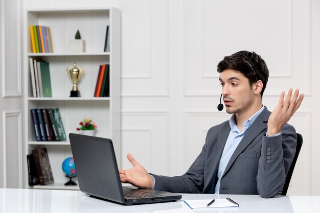 Service client mec mignon en costume gris avec ordinateur et casque agitant les mains lors d'un appel vidéo