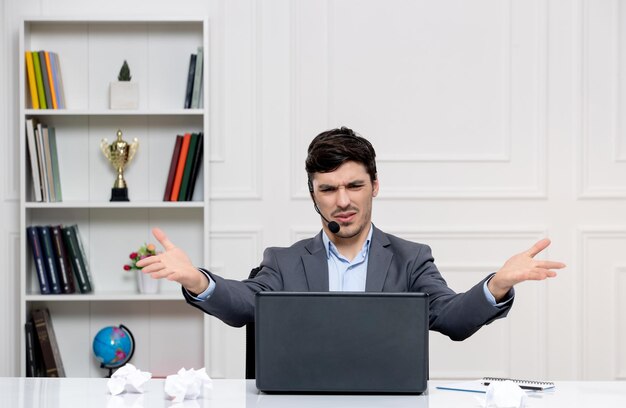 Service client mec mignon en costume gris avec ordinateur et casque avec du papier froissé sur le bureau