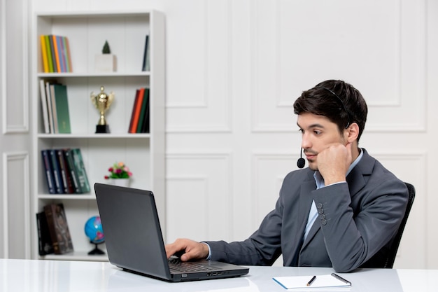 Service client mec mignon en costume gris avec ordinateur et casque avec la main sur le menton