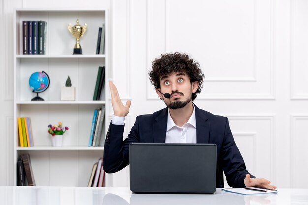 Service client mignon mec brune en costume de bureau avec ordinateur et casque pensant et levant les yeux