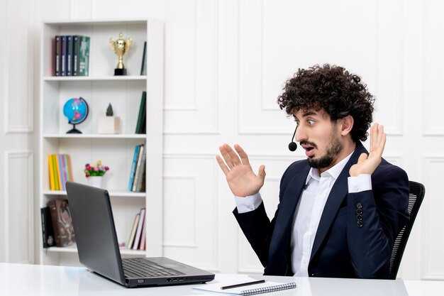 Service à la clientèle bel homme bouclé en costume de bureau avec ordinateur et casque agitant et choqué