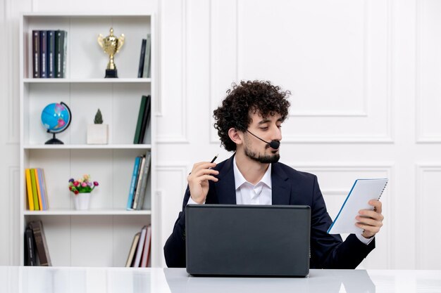 Service à la clientèle bel homme bouclé en costume de bureau avec ordinateur et casque lisant des notes