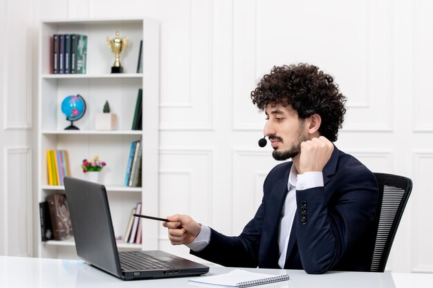 Service à la clientèle bel homme bouclé en costume de bureau avec ordinateur et casque pointant avec un stylo