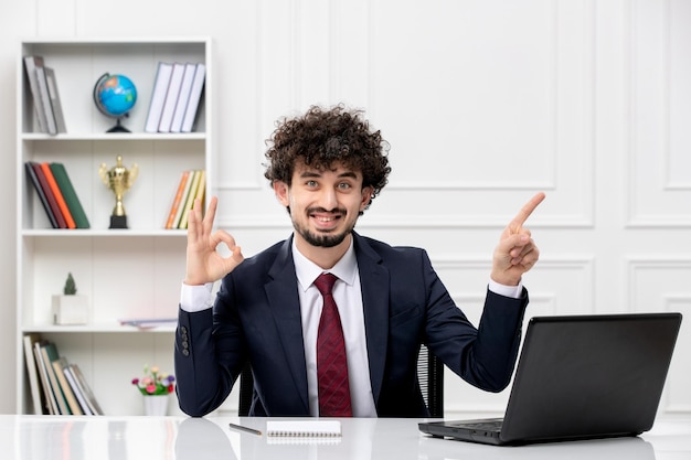 Service à la clientèle jeune homme brune bouclée en costume de bureau et cravate rouge avec ordinateur portable heureux et souriant