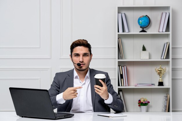 Service à la clientèle joli gentleman avec ordinateur en costume de bureau gris tenant une tasse de papier noir