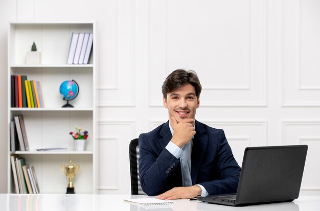 Service à la clientèle le joli mec brune en costume avec un ordinateur portable heureux de toucher le menton