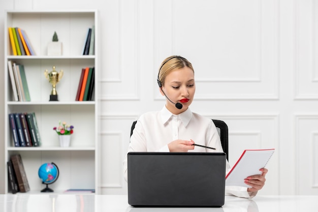 Service à la clientèle jolie fille blonde en chemise de bureau avec casque et notes de lecture d'ordinateur