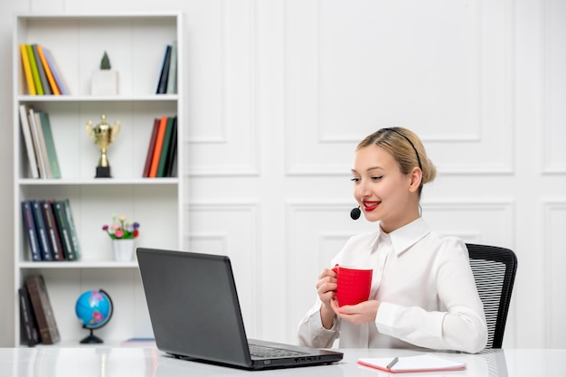 Service à la clientèle jolie fille blonde chemise de bureau avec casque et ordinateur tenant une tasse de café rouge