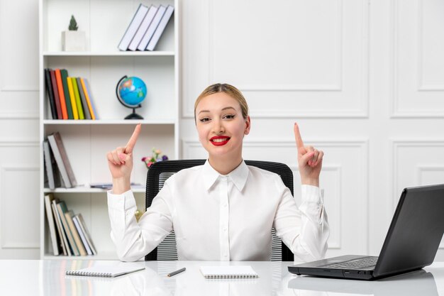 Service à la clientèle jolie fille en chemise blanche avec rouge à lèvres et ordinateur portable pointant vers le haut et souriant