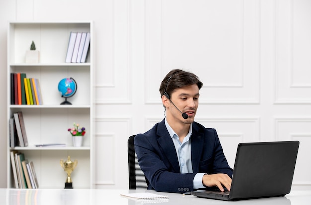 Service à la clientèle le mec mignon brune en costume avec un ordinateur portable et un casque dactylographie