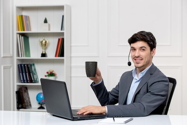 Service à la clientèle mec mignon en costume gris avec ordinateur et casque souriant avec une tasse de café