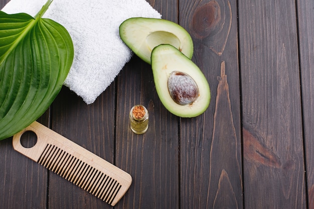 Serviette blanche, avocat, petite bouteille en verre et peigne en bois pour les cheveux se trouvent sur une table en bois