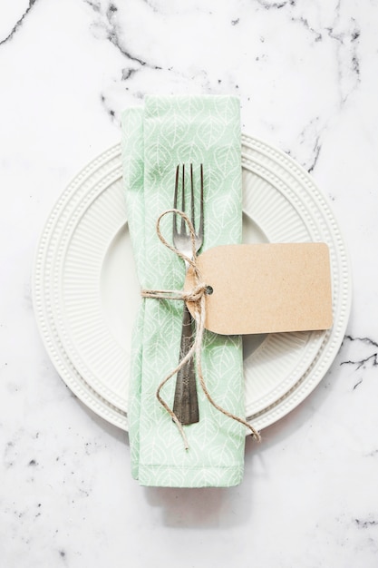 Serviette pliée et fourchette attachées avec une ficelle et une étiquette vierge sur une plaque en céramique blanche
