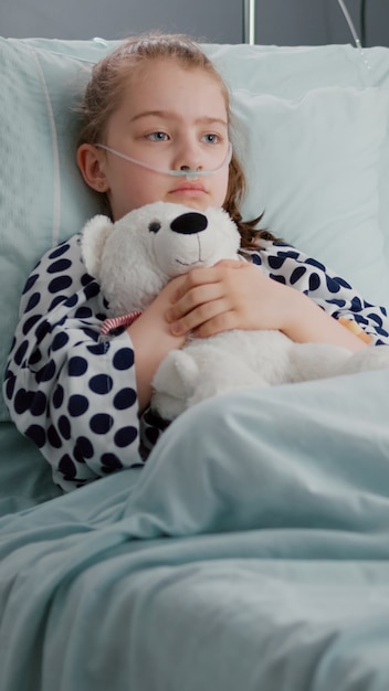 Photo gratuite seul enfant inquiet patient portant un tube nasal d'oxygène se reposant dans son lit tenant un ours en peluche