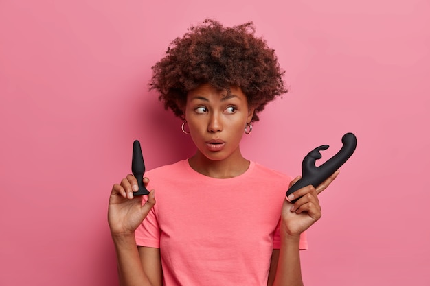 Sex toys et accessoires pour obtenir un plaisir érotique. Une femme surprise à la peau sombre regarde de côté, tient un vibromasseur noir et un plug anal pour l'intimité montre ce qu'elle a acheté dans un sex-shop. Femme avec des outils bdsm