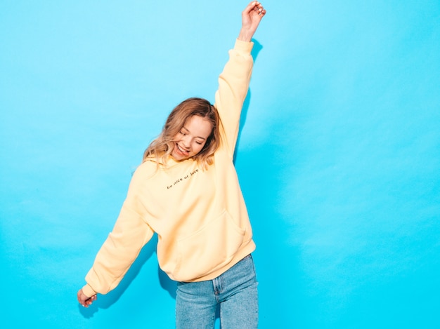 Sexy femme insouciante posant près du mur bleu. Modèle positif s'amusant, levant les mains et montrant la langue