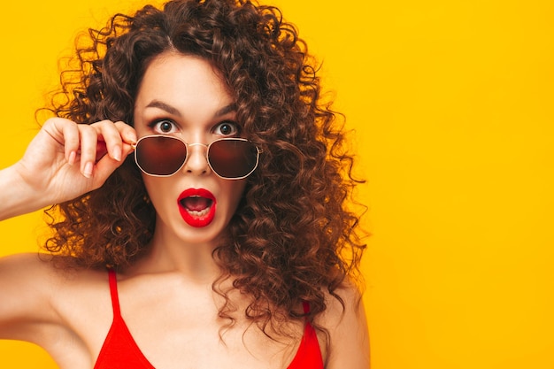 Sexy jeune belle femme souriante enlevant des lunettes de soleil femme insouciante isolée sur un mur jaune en studio Modèle brune positive avec une coiffure de boucles afro Choqué et surpris