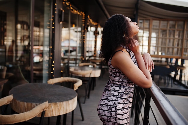 Sharming élégante jeune femme afro-américaine aux longs cheveux bouclés portant une combinaison posant à la terrasse du café en plein air contre la décoration de Noël Nouvel An