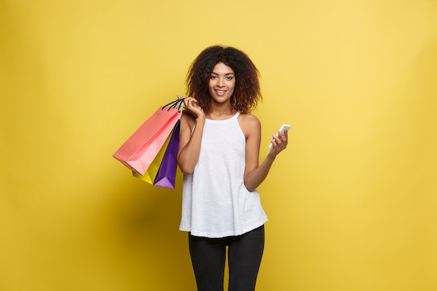 Photo gratuite shopping concept - close up portrait jeune belle attrayante femme africaine souriante et joyeuse avec sac à provisions coloré. fond en pastel pastel jaune. espace de copie.