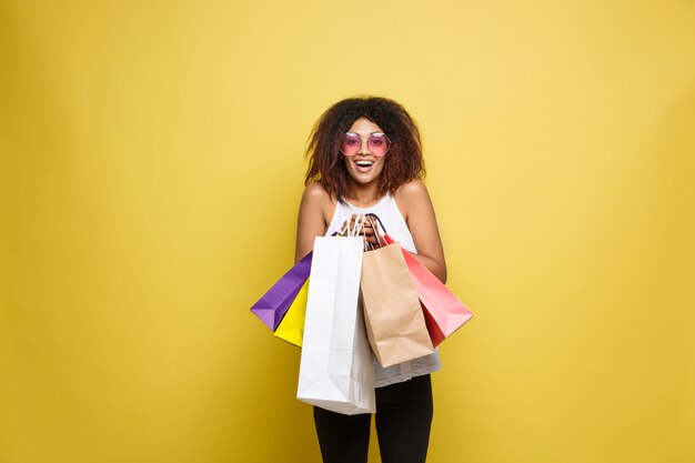 Shopping Concept - Close up Portrait jeune belle attrayante femme africaine souriante et joyeuse avec sac à provisions coloré. Fond en pastel pastel jaune. Espace de copie.