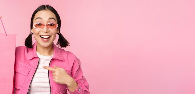 Shopping fille asiatique élégante à lunettes de soleil montrant le sac de la boutique et souriant recommandant la promotion de vente en magasin debout sur fond rose