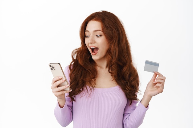 Shopping en ligne. Fille rousse excitée regardant l'écran du smartphone, lisant des réductions dans l'application, tenant une carte de crédit et un téléphone portable, debout sur fond blanc