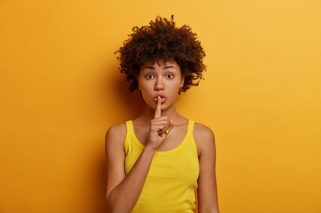 Silence et concept secret. Une femme afro-américaine surprise appuie son index sur ses lèvres, demande de se taire, raconte des informations très privées, porte un gilet jaune décontracté, pose, dit chut ou chut