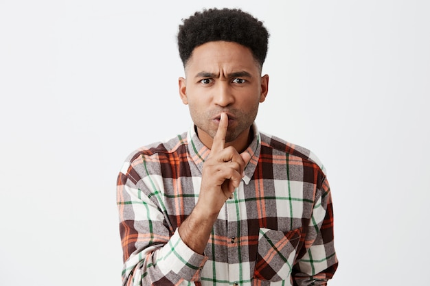 Silence. Gros plan de sérieux jeune beau homme à la peau sombre avec une coiffure afro en chemise à carreaux décontractée tenant le doigt devant la bouche, montrant le petit enfant à parler tranquillement dans les lieux publics.