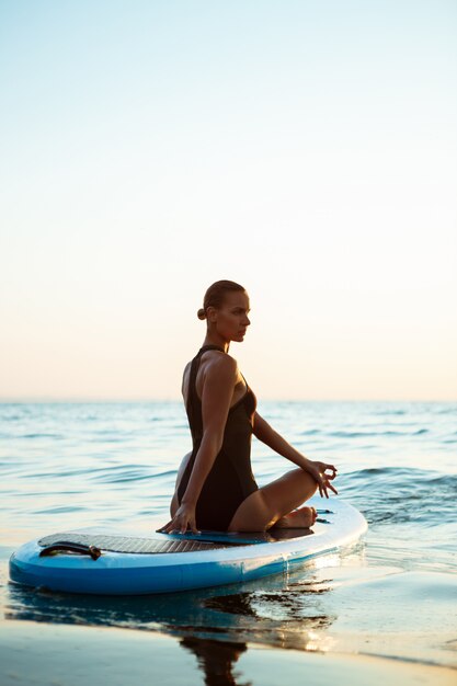 Silhouette de belle femme pratiquant le yoga sur planche de surf au lever du soleil.