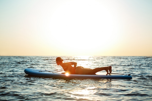 Photo gratuite silhouette de belle femme pratiquant le yoga sur planche de surf au lever du soleil.