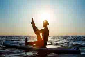 Photo gratuite silhouette de belle femme pratiquant le yoga sur planche de surf au lever du soleil.