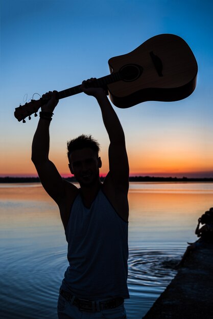 Silhouette de jeune bel homme tenant la guitare au bord de mer pendant le lever du soleil