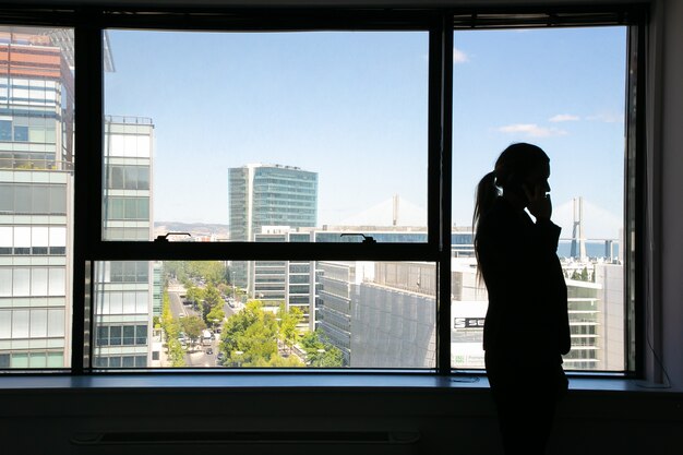 Silhouette méconnaissable de femme d'affaires debout près de la fenêtre et parler au téléphone mobile. Gestionnaire professionnel dans l'ombre et le paysage urbain. Concept d'entreprise, de communication et d'entreprise