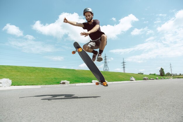 Skateur faisant un tour dans la rue de la ville en journée ensoleillée. Jeune homme en équipement d'équitation et de longboard près de prairie en action. Concept d'activité de loisirs, sport, extrême, passe-temps et mouvement.