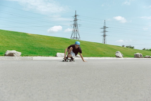 Skateur faisant un tour dans la rue de la ville en journée ensoleillée. Jeune homme en équipement d'équitation et de longboard près de prairie en action. Concept d'activité de loisirs, sport, extrême, passe-temps et mouvement.