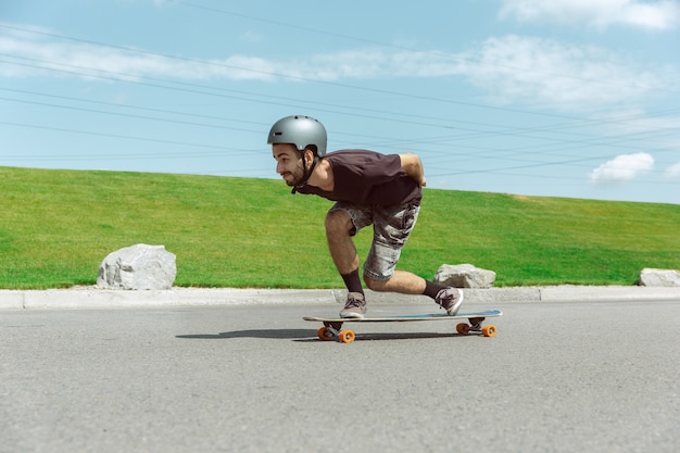 Photo gratuite skateur faisant un tour dans la rue de la ville en journée ensoleillée. jeune homme en équipement d'équitation et de longboard près de prairie en action. concept d'activité de loisirs, sport, extrême, passe-temps et mouvement.