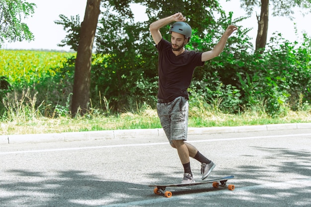 Skateur faisant un tour près de la prairie en journée ensoleillée. Jeune homme en équipement d'équitation et de longboard sur l'asphalte en action. Concept d'activité de loisirs, sport, extrême, passe-temps et mouvement.