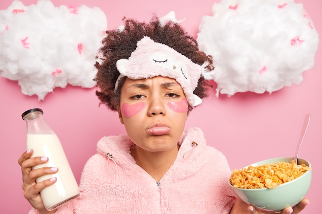 Sleepy bouder femme afro-américaine avec une expression triste porte des vêtements de nuit mange des aliments sains applique des patchs de collagène sous les yeux pose contre le mur rose