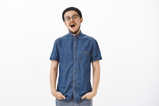 Sleepy fatigué beau mec avec barbe à lunettes et bonnet noir bâillant et plissant les yeux voulant dormir sur un mur gris