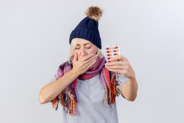 Sleepy young blonde femme slave malade portant un chapeau d'hiver et une écharpe bâille mettant la main sur la bouche et détient pack de pilules médicales isolé sur un mur blanc avec copie espace