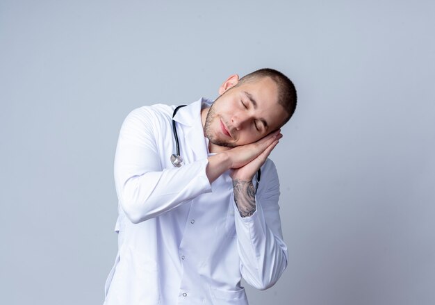 Sleepy young male doctor portant une robe médicale et un stéthoscope autour de son cou faisant le geste de sommeil isolé sur un mur blanc