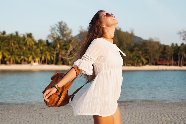 Slim belle femme en robe blanche sur la plage tropicale au coucher du soleil tenant un sac à dos en cuir.