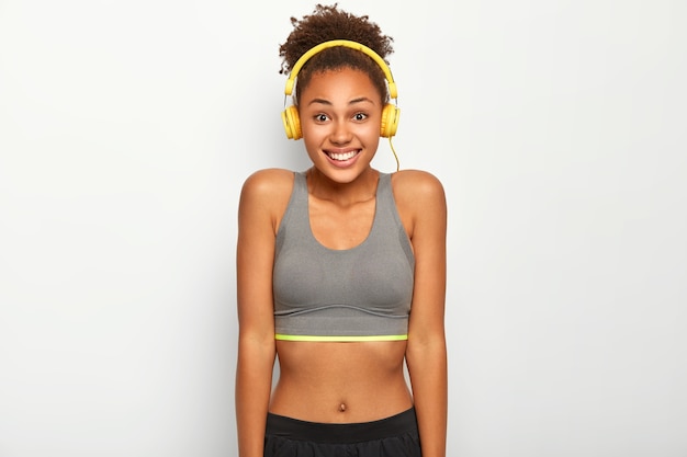 Photo gratuite slim femme afro sportive sourit joyeusement à l'avant porte haut décontracté, écouteurs sur les oreilles, écoute la musique préférée pendant l'entraînement